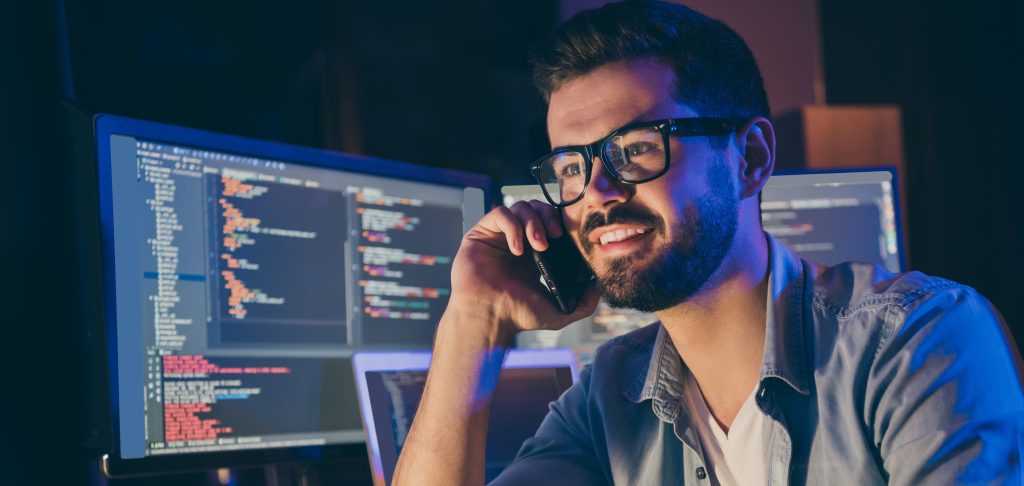 Close-up portrait of his he nice attractive skilled cheerful cheery guy geek tech shark talking on phone with friend remote help assistance service in dark room workplace workstation indoors